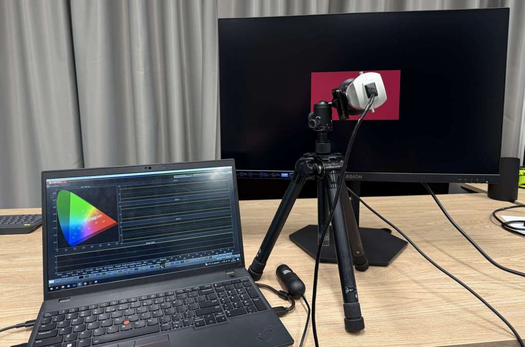 Monitor Lenovo Legion 27Q-10 na biurku podczas testu, obok urządzenie pomiarowe i laptop